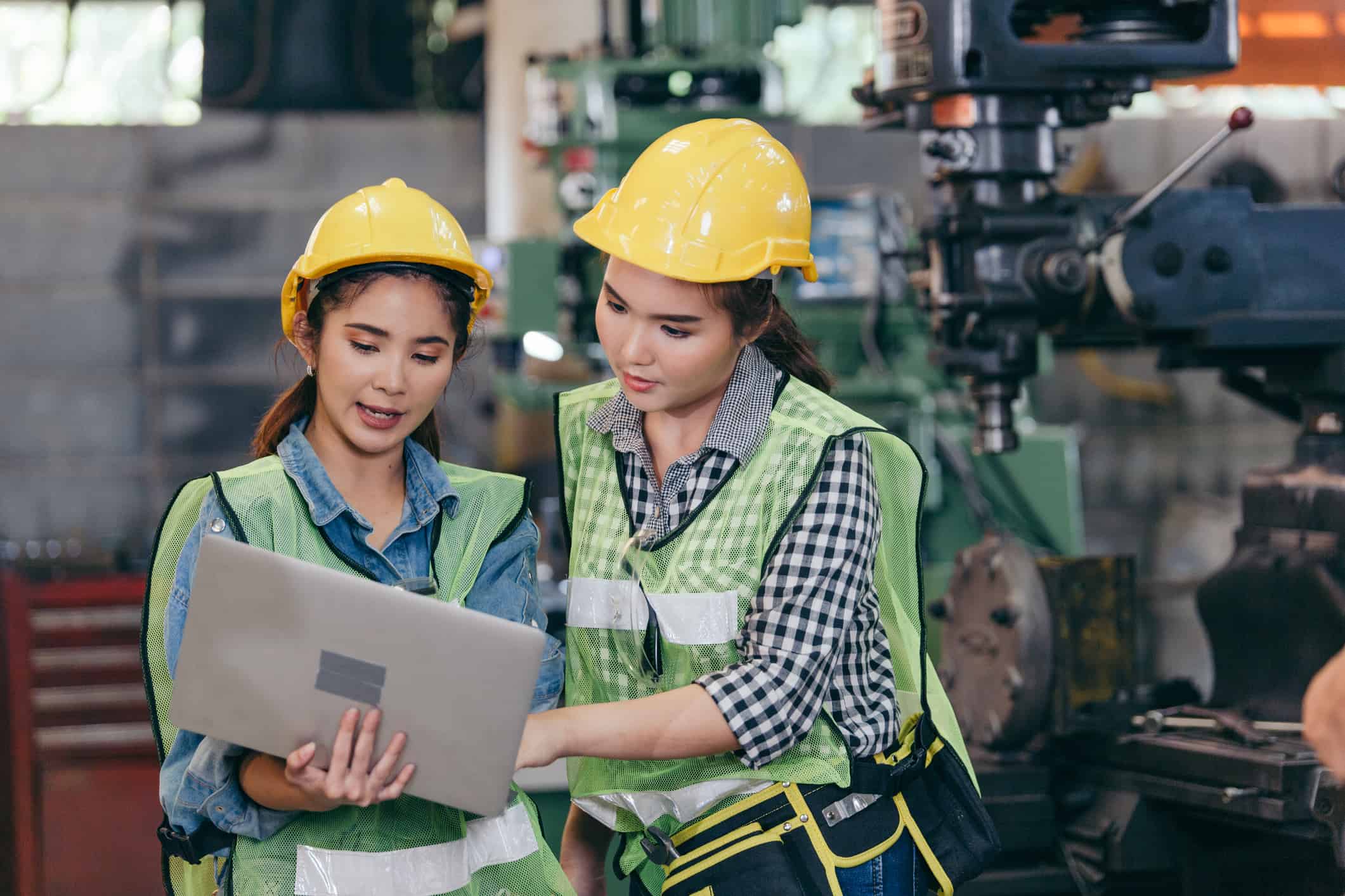 Two Asian female engineer workers wearing safety helmet discuss project in industry manufacturing. industrial woman watching laptop checking machine factory with team colleague. successful teamwork